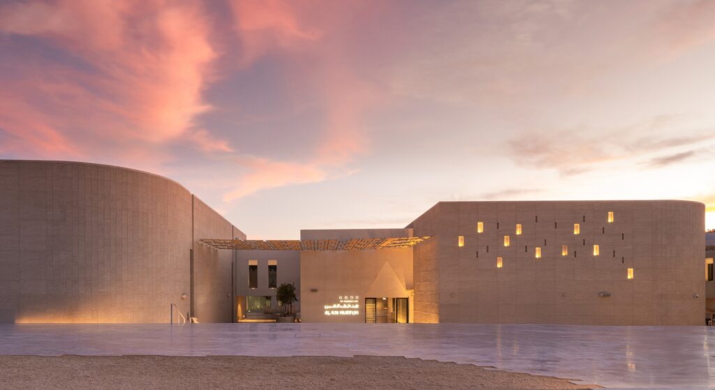 Main entrance of Al Ain Museum under a sunset sky with glowing wall niches and modern Arabic calligraphy signage.