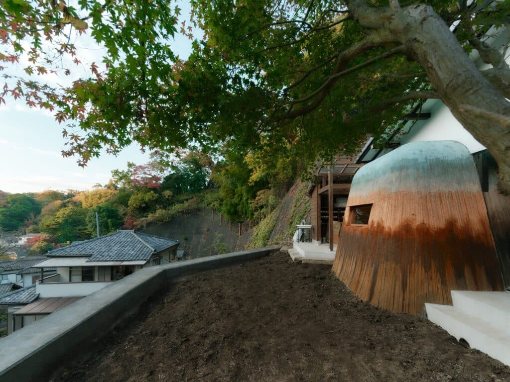 Wide exterior shot of Haniyasu House on a sloped site in Kamakura, showing the building&rsquo;s integration with the steep landscape and trees.