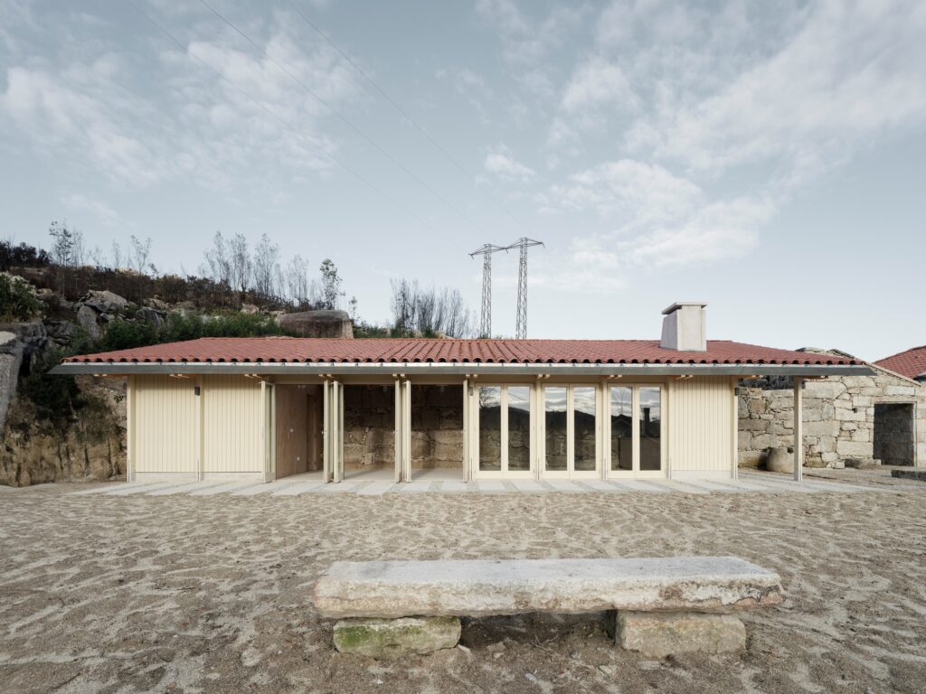 Front view of the elongated wooden pavilion with large glass doors opening onto a sandy courtyard with a stone bench.