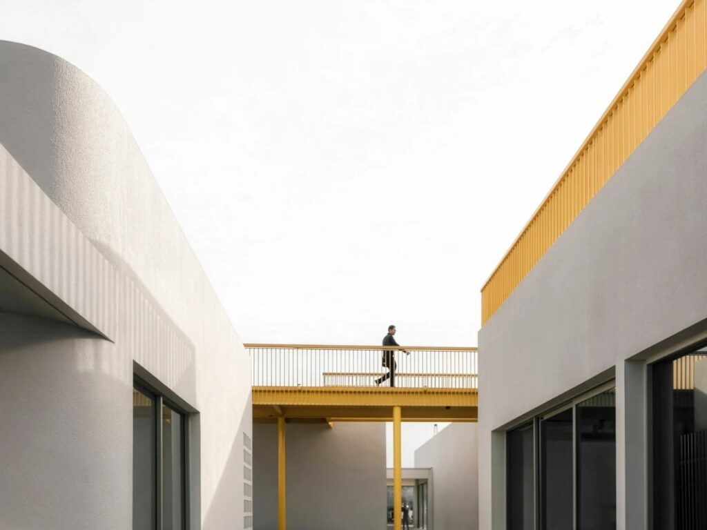 A person walking on a yellow bridge between minimalist white architectural structures under a clear sky.