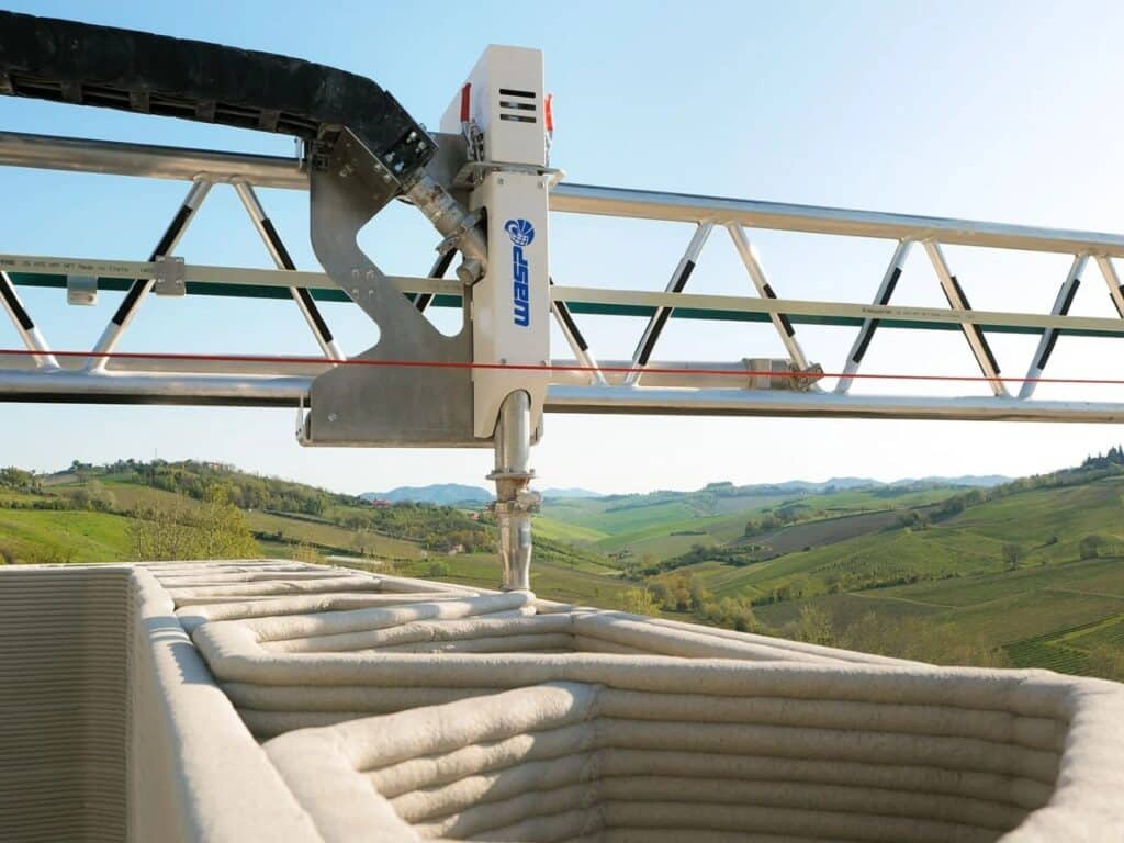Extreme close-up of a 3D printing nozzle depositing material on a hollow wall structure with a scenic valley in the background.