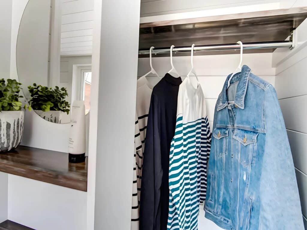 A compact integrated closet inside the Kinnakeet house with hanging clothes next to a bathroom mirror and vanity.