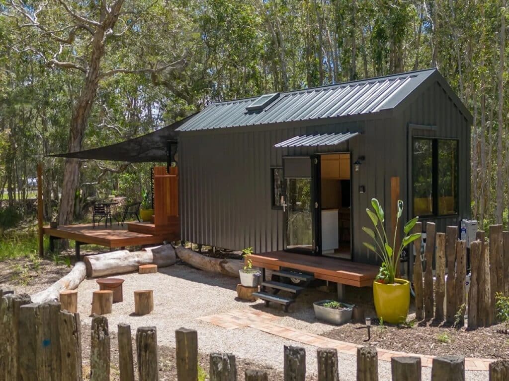 Full exterior shot of the Artista tiny house on wheels, showcasing its dark corrugated metal cladding and sustainable design in a forest setting.