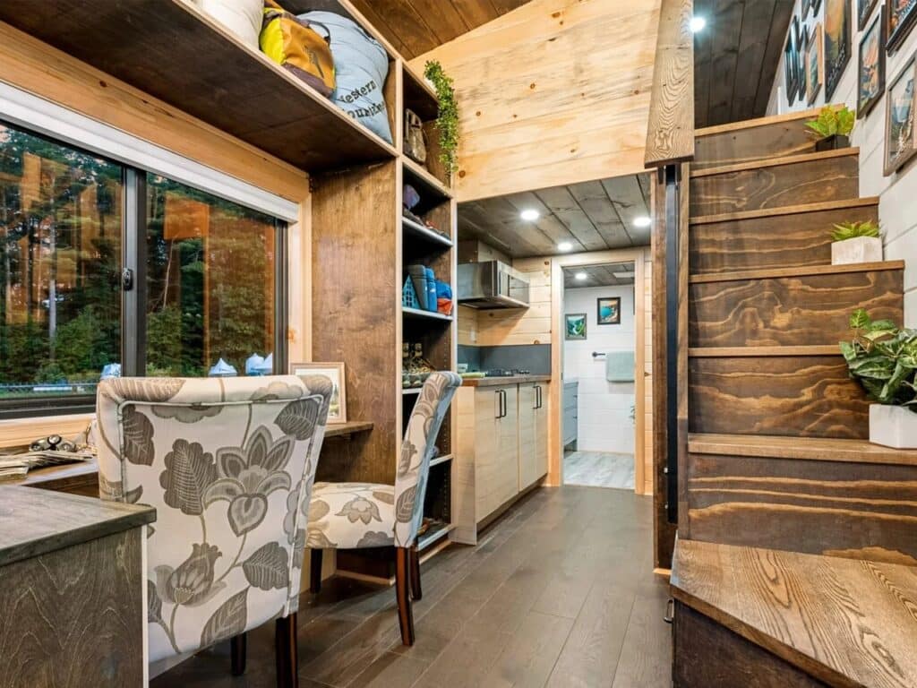 Interior view of a tiny home workspace with wooden stairs, storage shelves, and a view into the kitchen.