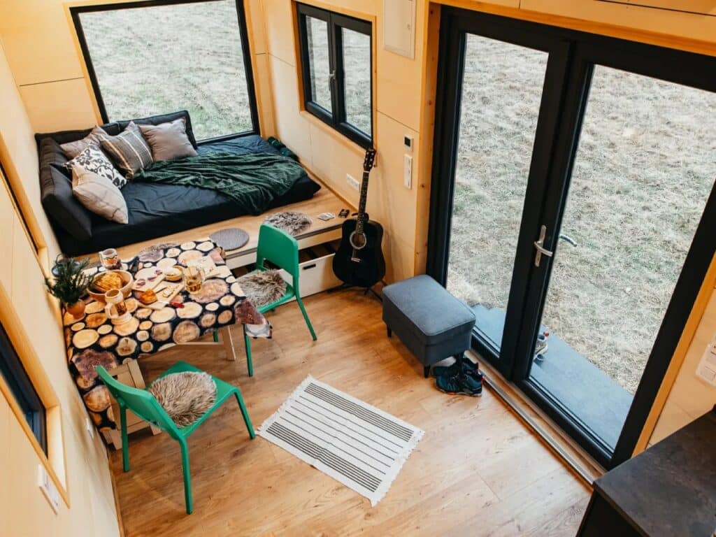 High-angle interior view of a tiny house living area with a cozy daybed, multifunctional dining table, and large glass doors opening to nature.