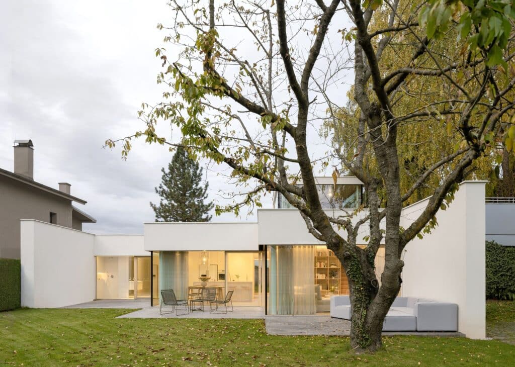 Evening view of Sestajovice House showing illuminated interior spaces through large glass openings and a central tree.