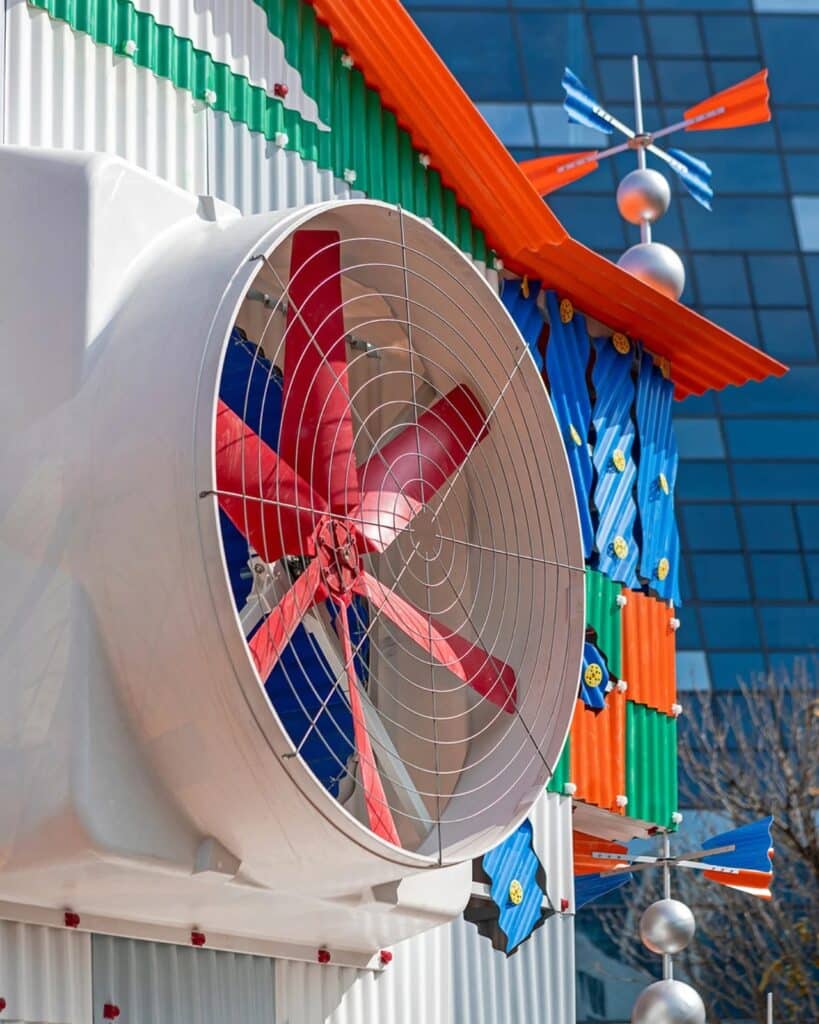 Angled side view of Clock House No. 2 showing a large fan unit and a silver weather vane against a clear blue sky.