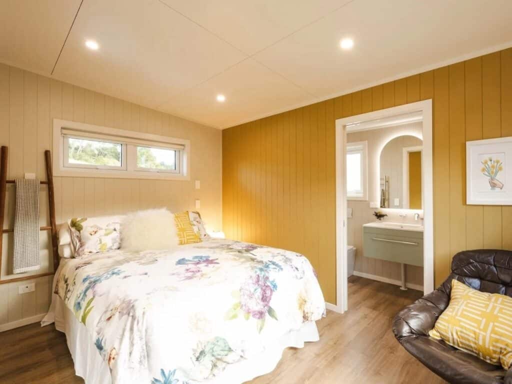 Tiny house bedroom featuring floral bedding, a mustard yellow accent wall, and a direct view into the ensuite bathroom.