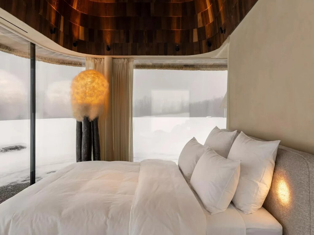 Interior of a modern bedroom inside the Mazanka house featuring a panoramic glass wall overlooking a snowy field and a warm illuminated cloud lamp.