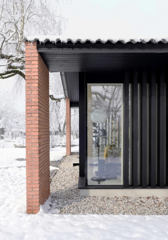 Side view of the house showing vertical black wooden slats and a brick pillar in the snow.