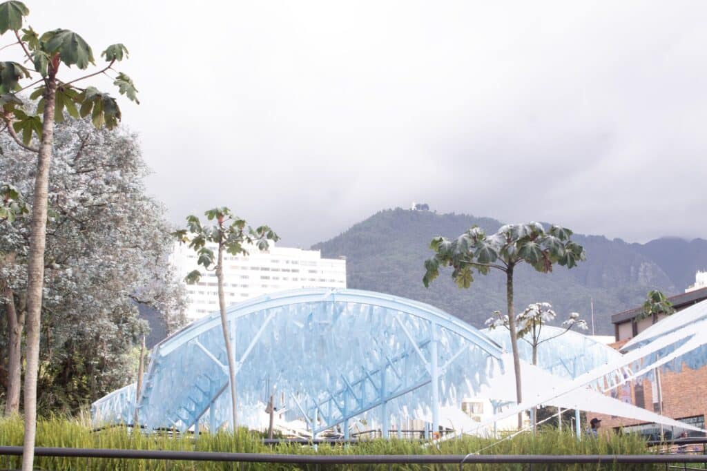 Low-angle shot of the light blue arched pavilion structure against a cloudy sky and distant hills, with tropical trees in the foreground.