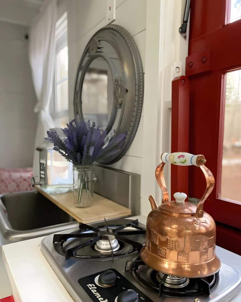 Close-up of a compact kitchen in a tiny house with a copper kettle on a two-burner stove and a round mirror.