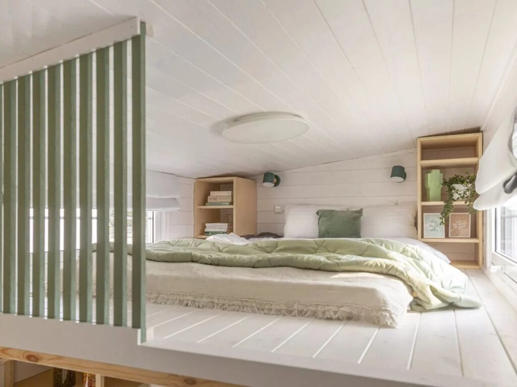 A loft-style bedroom in a tiny house with a double bed, wooden side shelves, and white wood-paneled walls.