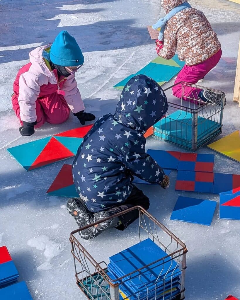 Children in winter gear playing with large colorful geometric tiles on the ice, participating in an art project.