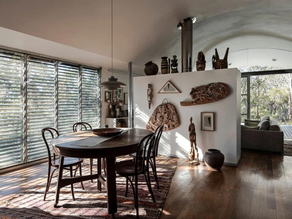 Dining area with a wooden table, louvered windows for ventilation, and a wall displaying indigenous sculptures.