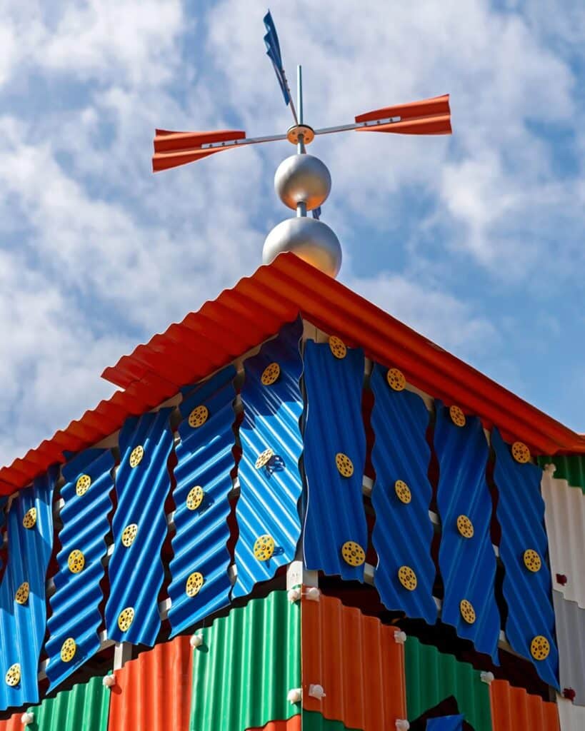 Low angle view of the roof corner of Clock House No. 2 with blue corrugated panels and a weather vane pointing toward the clouds.