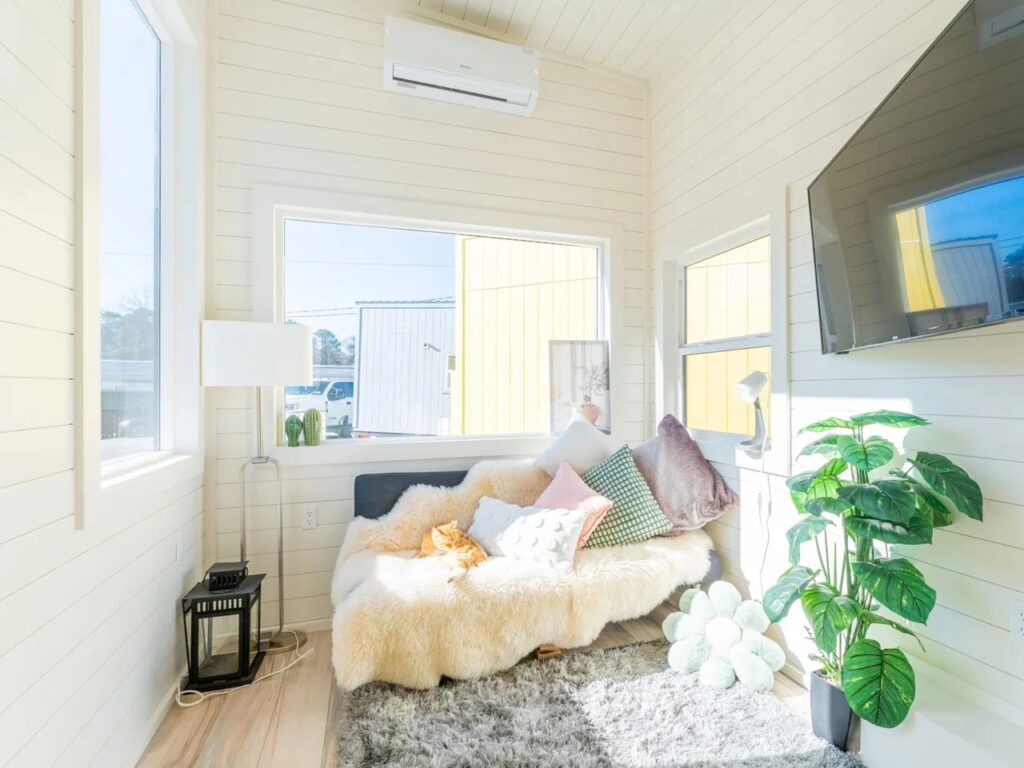 Cozy living area in a tiny house with large windows, natural light, and a comfortable sofa under an AC unit.