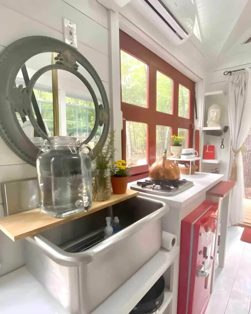 Wide shot of the tiny house kitchen area with white walls, red window frames, and a stainless steel farmhouse sink.