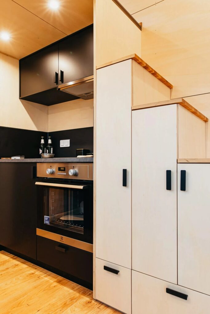 Built-in kitchen storage and appliances in a tiny house, featuring a modern oven and white minimalist cabinets under a loft staircase.