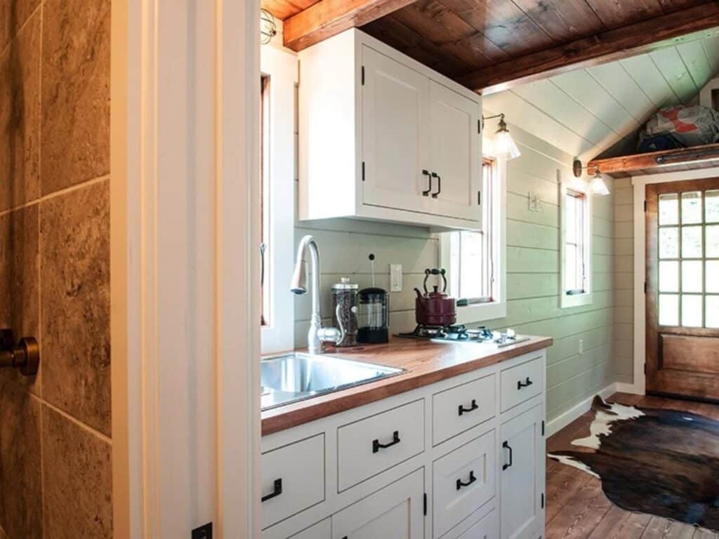 Detailed view of a small kitchen in a tiny house with white cabinets, wood countertops, a stainless steel sink, and a red kettle.
