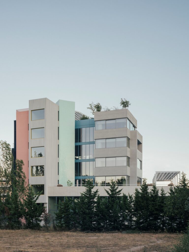 A full view of the rehabilitated administrative building featuring tiered levels, colorful vertical sections, and integrated greenery behind a row of pine trees.