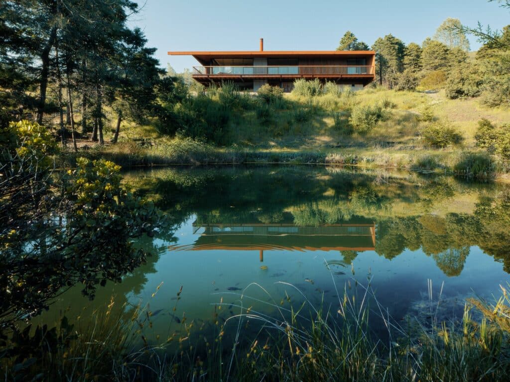 Reflection of the modern steel house in a still natural water pond surrounded by trees.