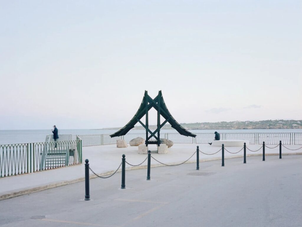 Distant view of the Asympta structure on a pier, showing its symmetrical A-frame silhouette and a person sitting nearby for scale.