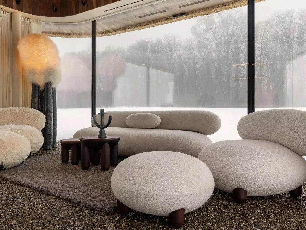 Living room area with rounded white textured sofas, pebble flooring, and a large window looking out to a misty forest.