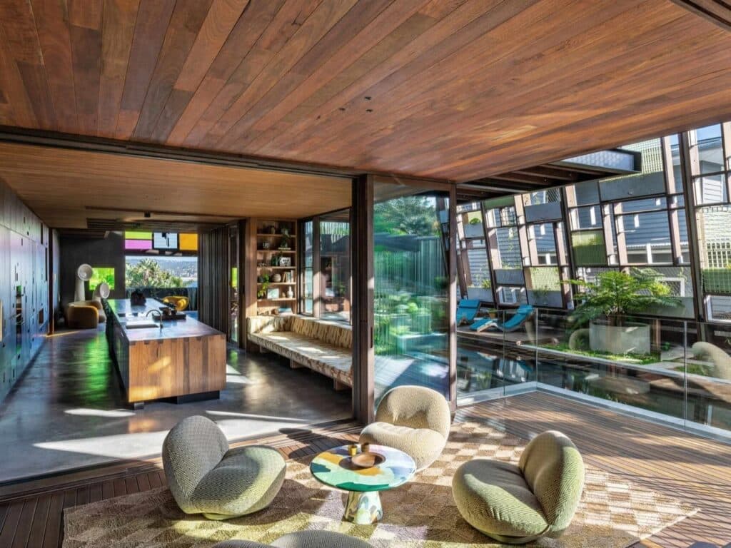 Wide view of an open-plan living area in Holocene House showing a long wooden kitchen island, a natural pool, and a multi-layered ceiling.