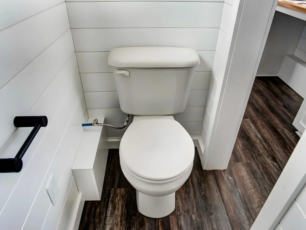 A clean, white minimalist bathroom in a tiny house featuring a standard toilet and horizontal wood paneling.