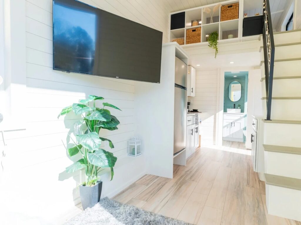 View from the living area towards the bathroom and loft stairs in the Sora 20 tiny home, featuring a wall-mounted TV.