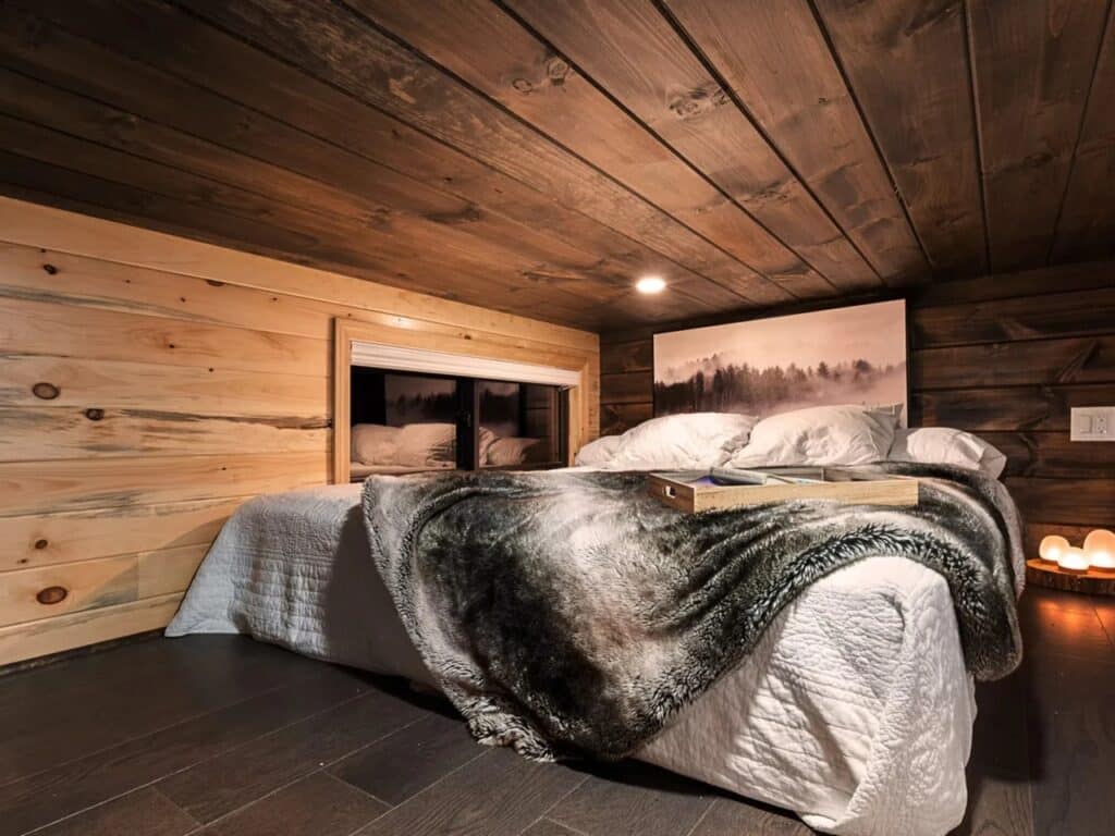 Loft bedroom in a tiny home with dark wood ceilings, a cozy bed, and warm ambient lighting.