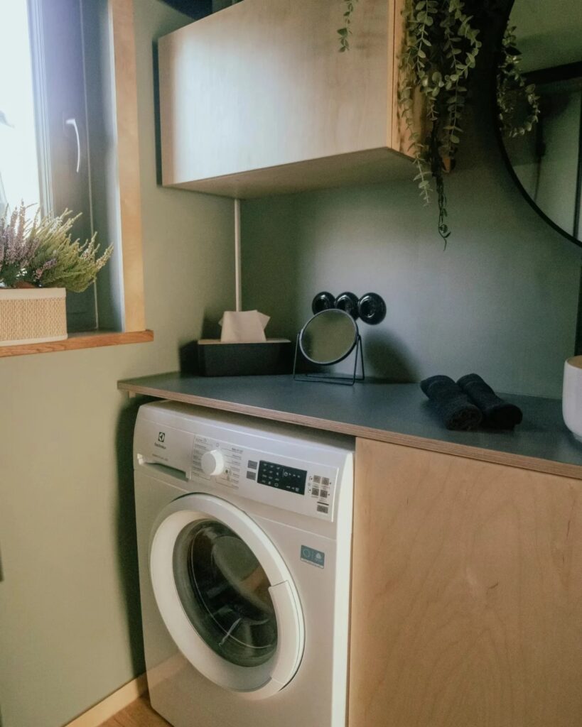 Modern tiny house bathroom corner featuring a washing machine integrated under a wooden countertop.