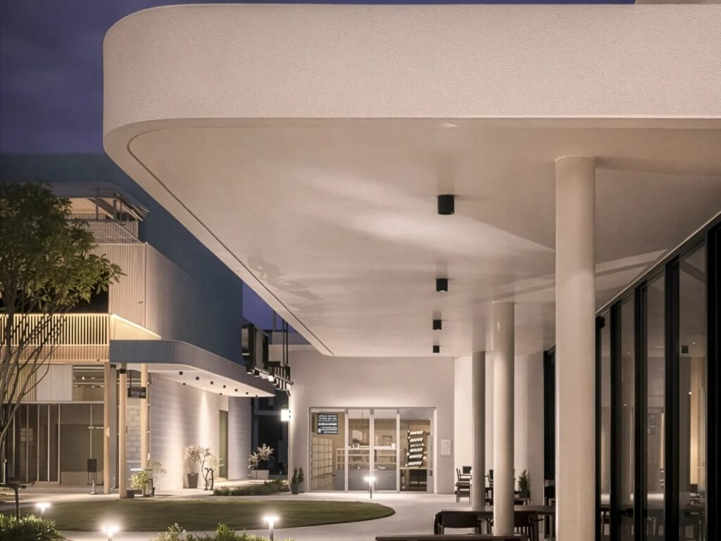 Modern white building facade with a deep overhang and warm interior lighting at dusk.