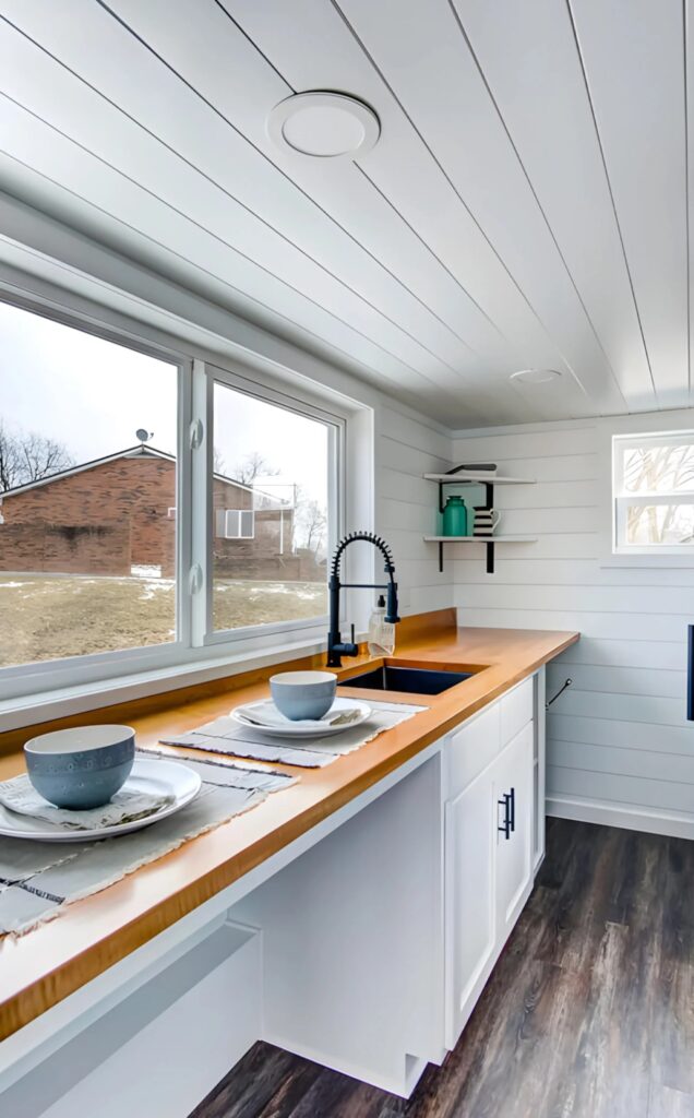 A narrow functional kitchen with a long wooden countertop, white cabinets, and a modern black faucet overlooking a window.