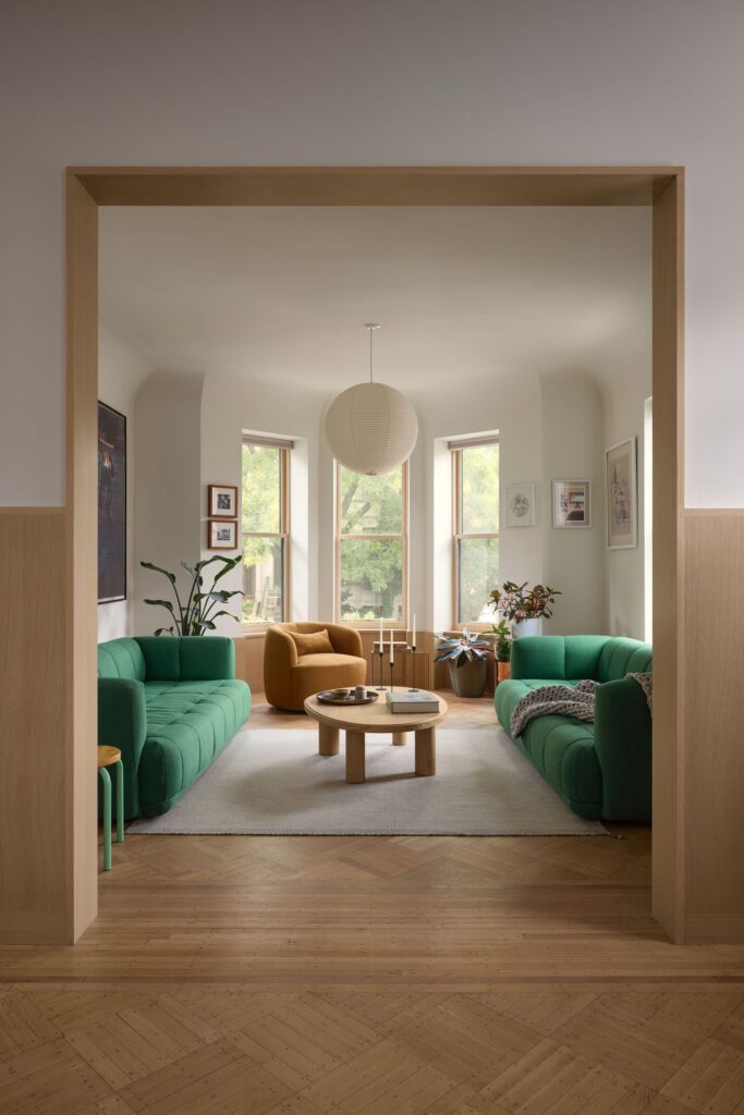 View through a wide wooden framed doorway into a sunlit living room with green seating and bay windows.