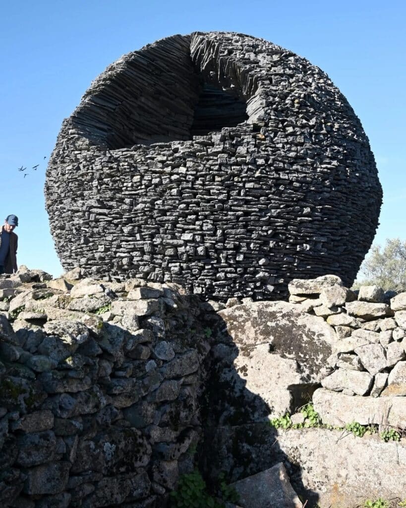 The stone sphere seen from behind a low stone wall, with birds flying in the background against a blue sky.