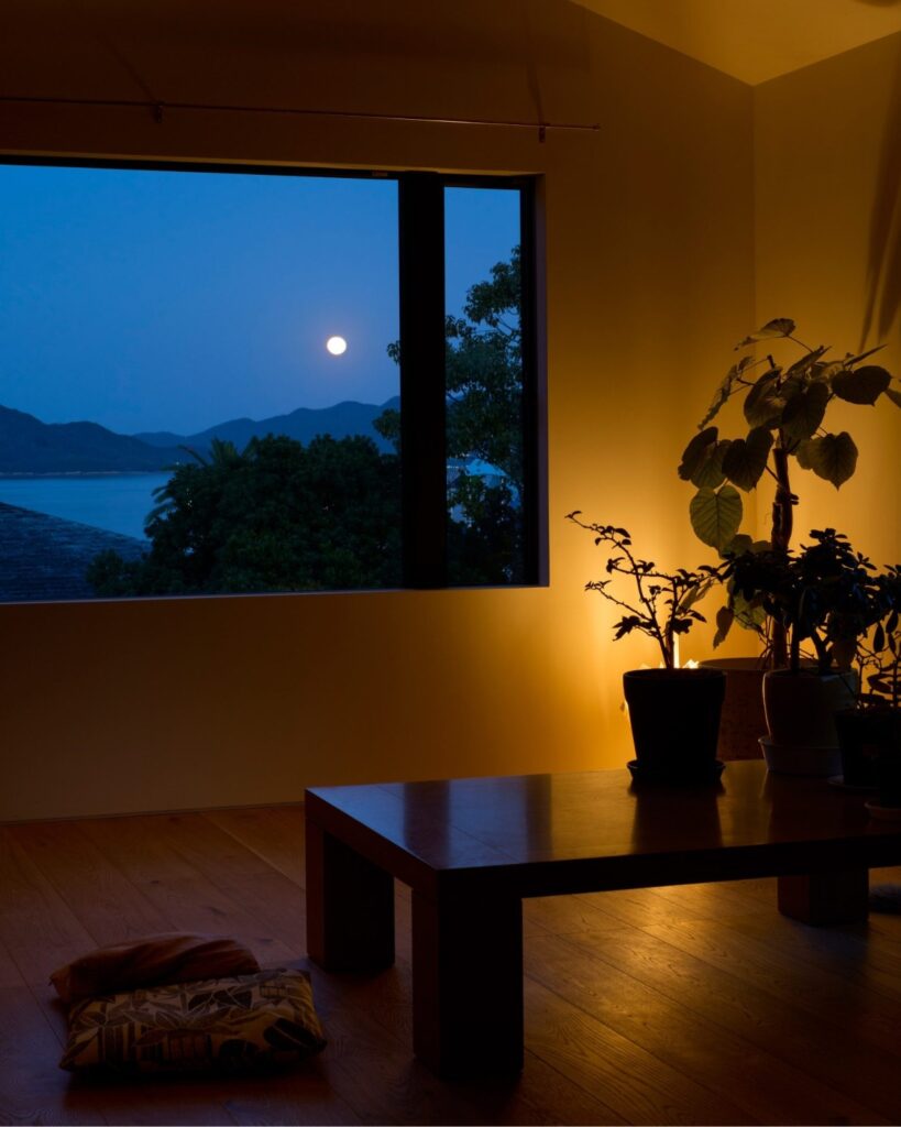 Interior view of Onomichi House living space at night, featuring a large window with a view of the sea and mountains under moonlight.