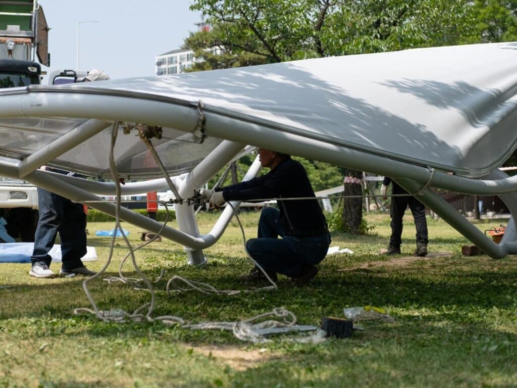 Construction workers installing a Seoul Wing canopy using ropes and light machinery in a park.