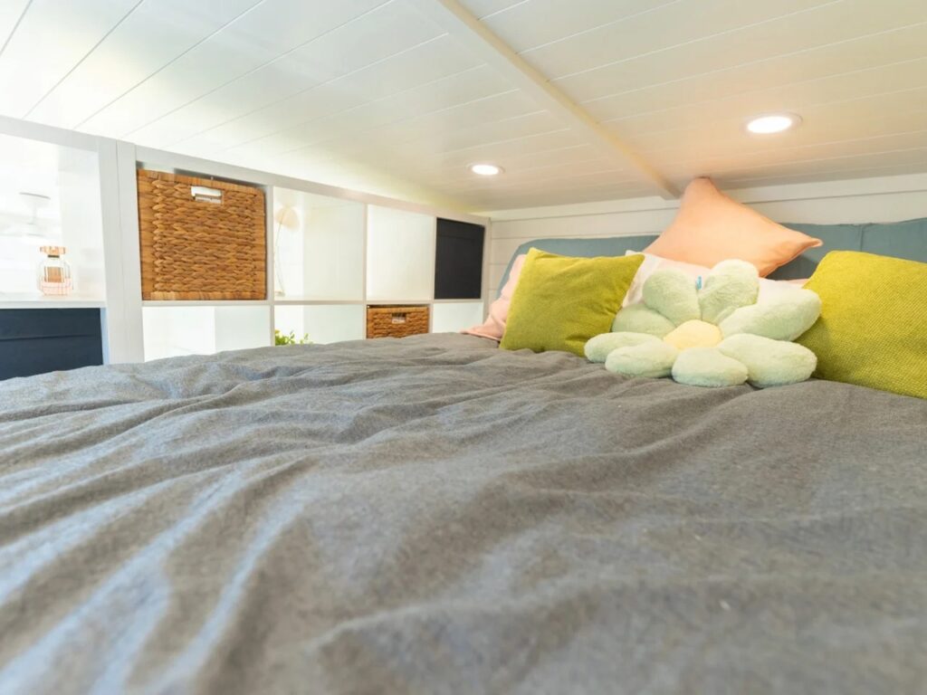Close-up of the upper sleeping loft in the Sora 20 tiny house with integrated shelving and gray bedding.