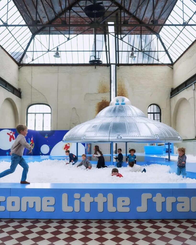 Children playing and running in a white ball pit around a silver UFO structure in a high-ceiling industrial space.