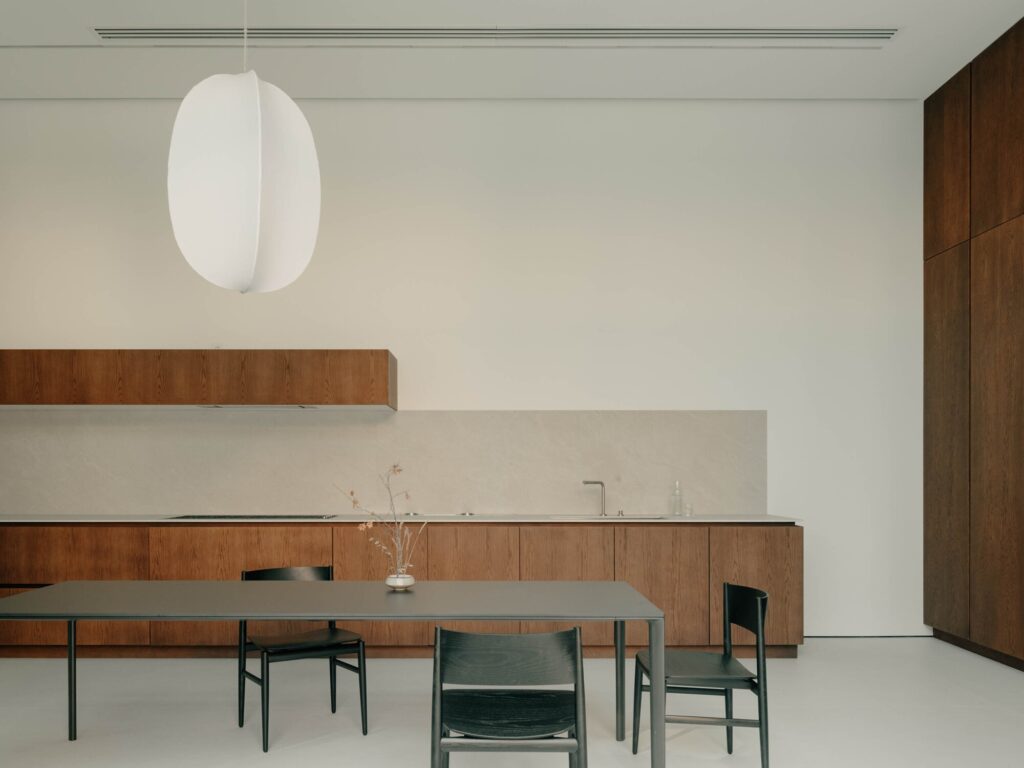 Minimalist dining table and wooden kitchen cabinetry under a soft white pendant lamp.