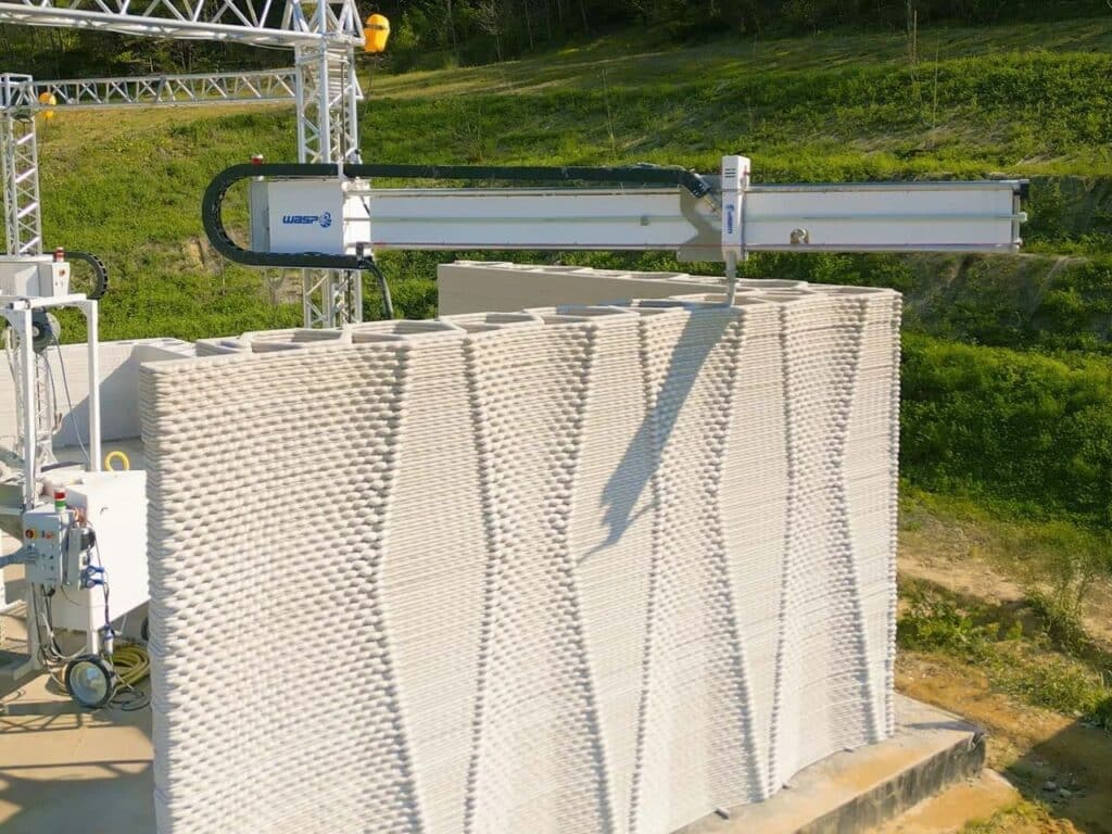 A WASP 3D printing arm in action, finishing the top layers of a tall, textured wall in a rural Italian setting.