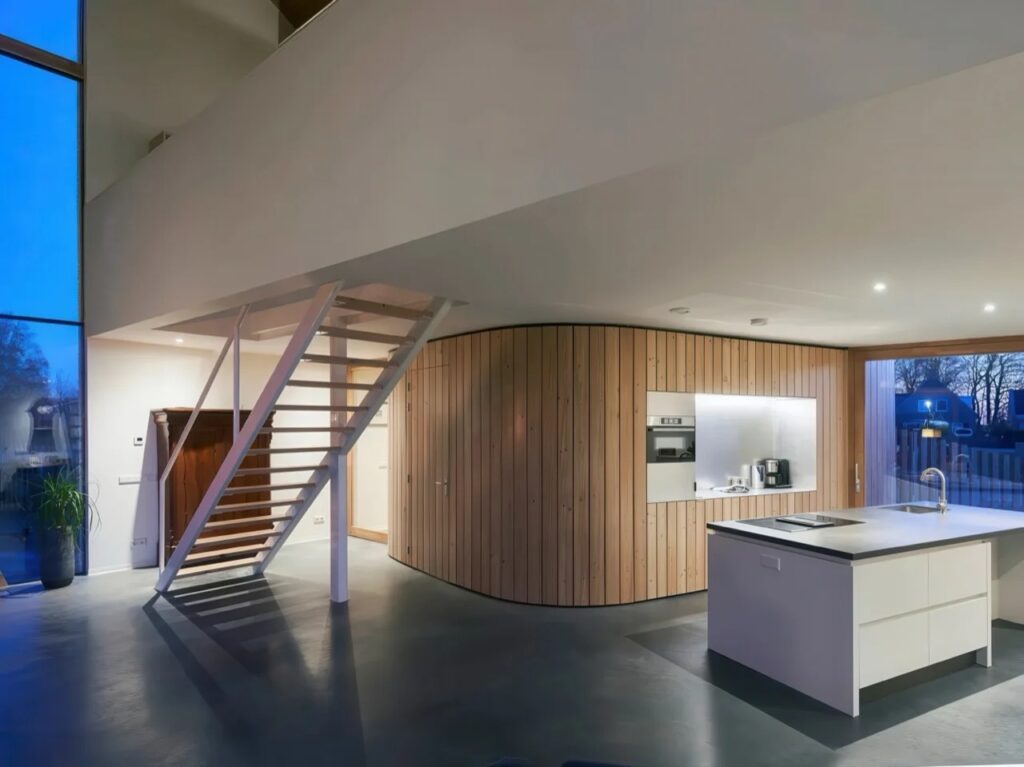 Modern kitchen at night featuring a curved timber wall, white island counter, and polished concrete flooring.