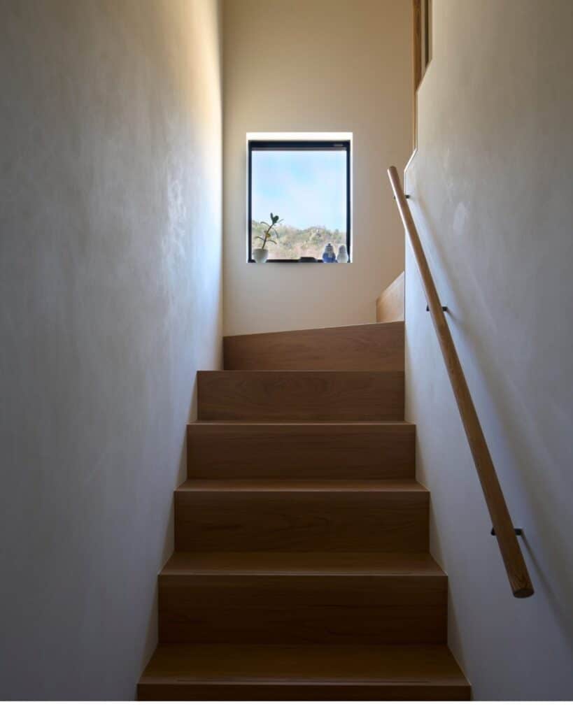 Minimalist wooden staircase inside Onomichi House, illuminated by natural light from a small square window at the top.