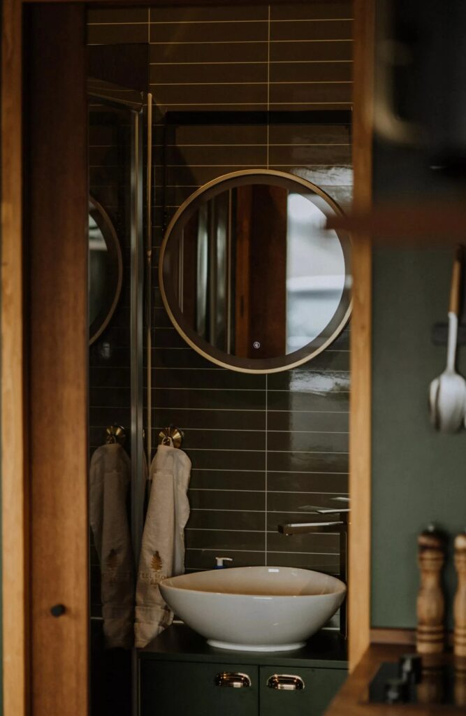 Bathroom interior featuring a round backlit mirror, a vessel sink, and dark tiled walls.