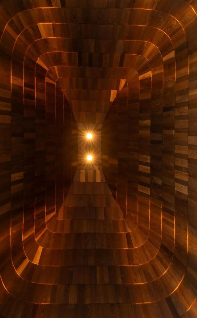 Looking up into the high vaulted ceiling made of dark wood shingles with warm glowing lights at the apex.