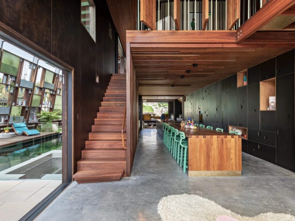 Contemporary home interior with a dramatic wooden staircase, high ceilings, and a long green-stooled kitchen island next to a pool view.