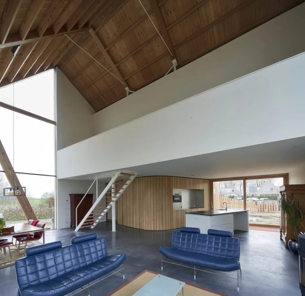 Wide-angle interior view of the open-plan house with blue sofas, a mezzanine level, and a high timber-framed ceiling.