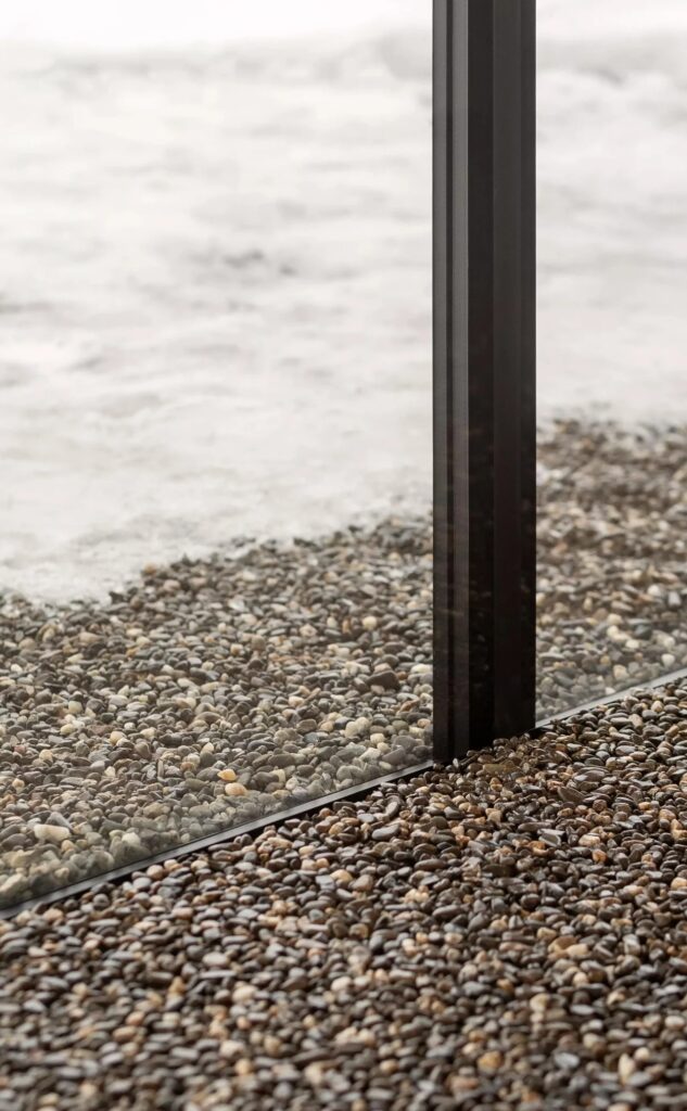 Detail of the glass wall meeting a floor made of small natural river pebbles, with snow visible outside.